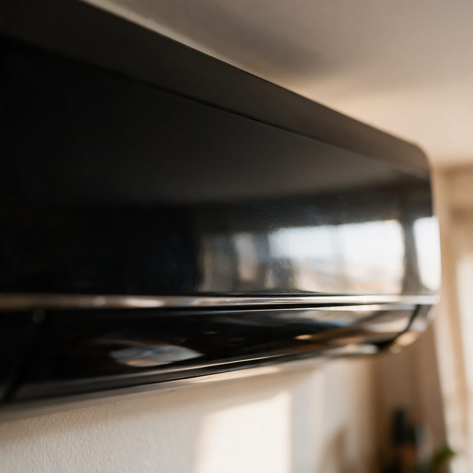 A glossy black indoor air-conditioner panel is shown close up at an angle, mounted near a ceiling in a warm-lit room with soft daylight and a wall visible below.