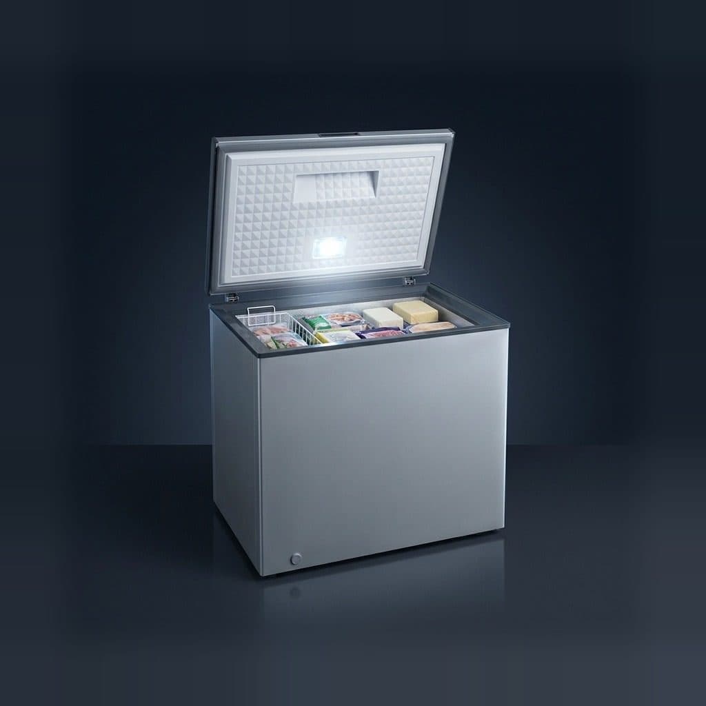 A silver single-lid chest freezer stands open on a dark reflective studio floor against a navy back-wall gradient, its lid lamp lighting the quilted liner, a wire basket on the rim and stacked frozen packs.