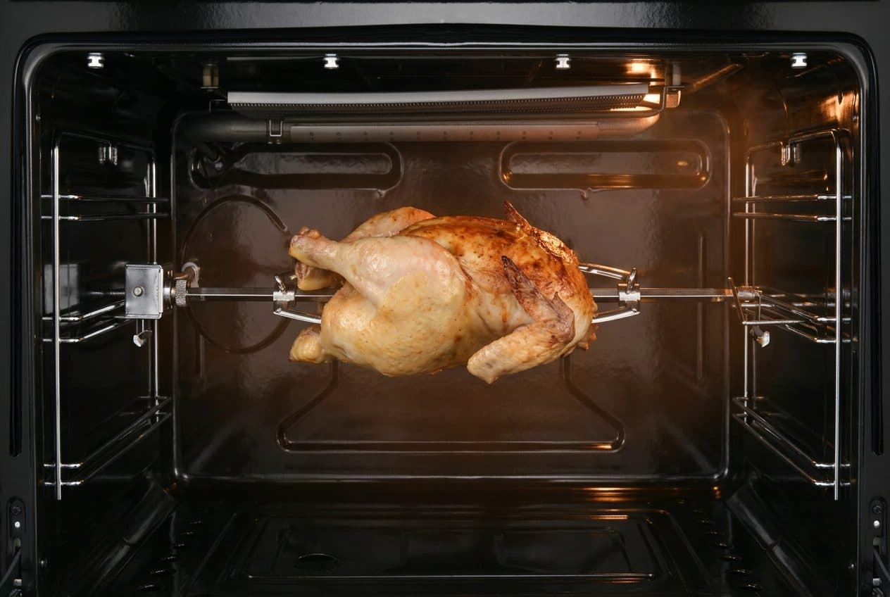A whole roasted chicken turns on a rotisserie spit inside a lit, open gas-oven cavity, its skin golden-browned, with chrome shelf runners and a radiant grill element visible along the top of the black enamel interior.