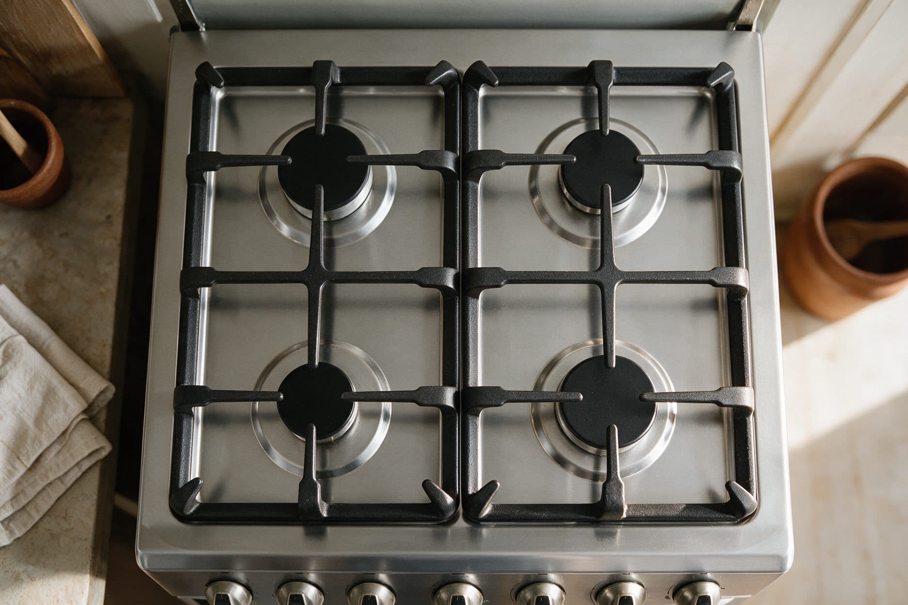 A stainless steel four-burner gas hob sits on a kitchen counter in warm natural light, with a linen cloth, wooden utensil holder and terracotta pots at the edges of frame.