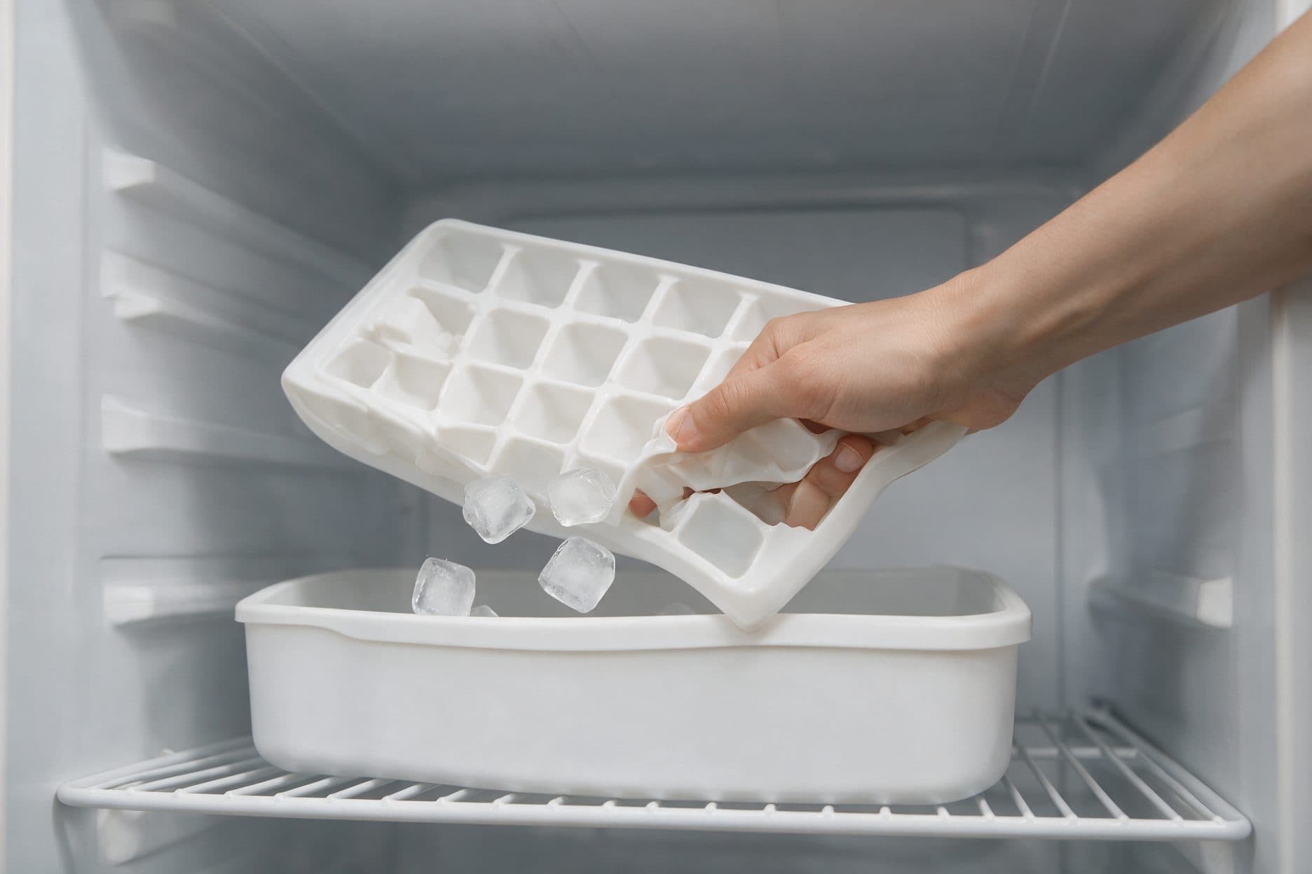 A hand twists a white plastic ice tray over a refrigerator's open container, with cubes dropping free.