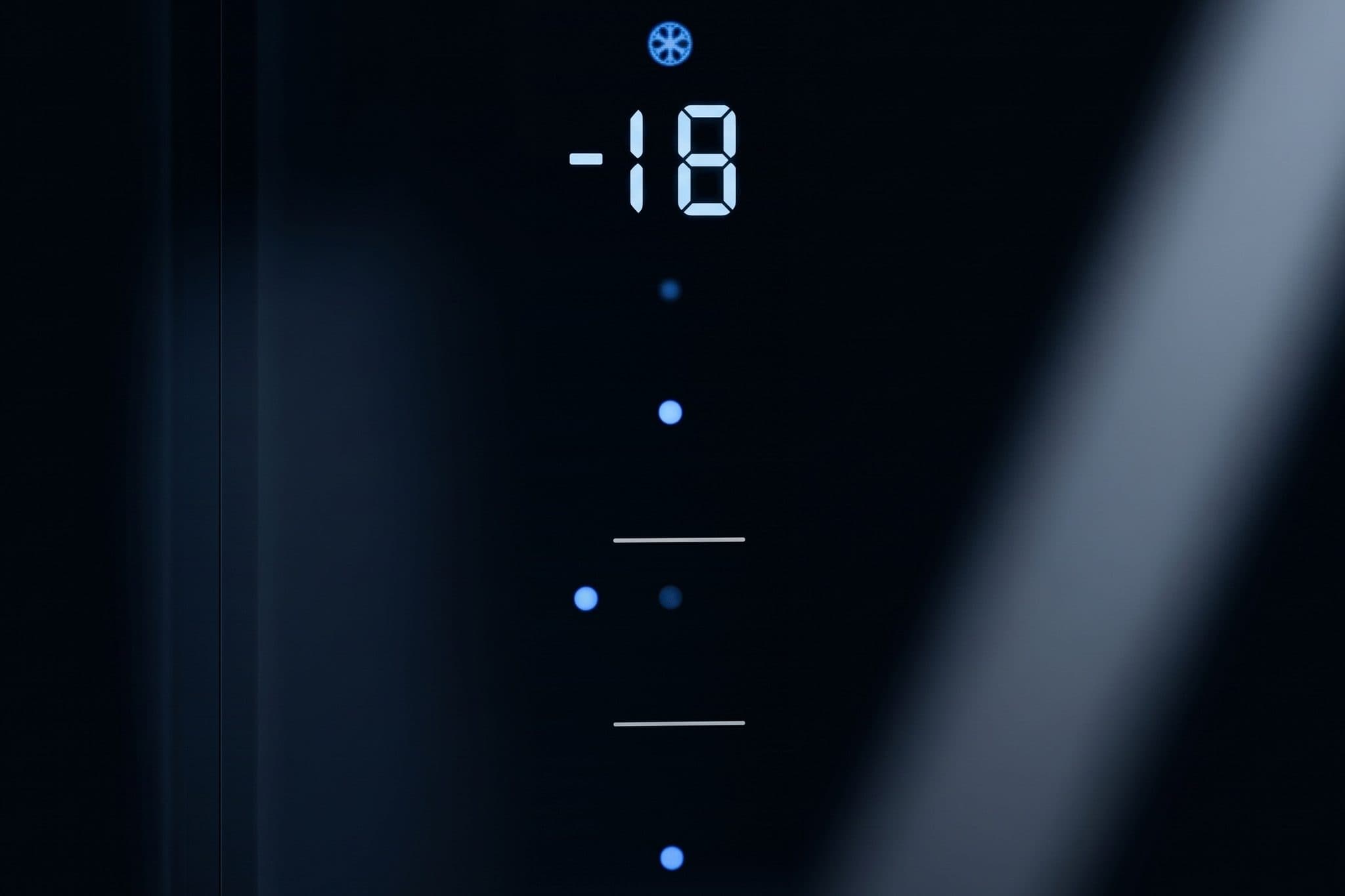 Ice-blue seven-segment digits reading -18 glow under a small snowflake icon, above a column of small blue dots and two thin bars, on a black glass refrigerator door, with a hairline door seam to the left and a soft diagonal reflection to the right.