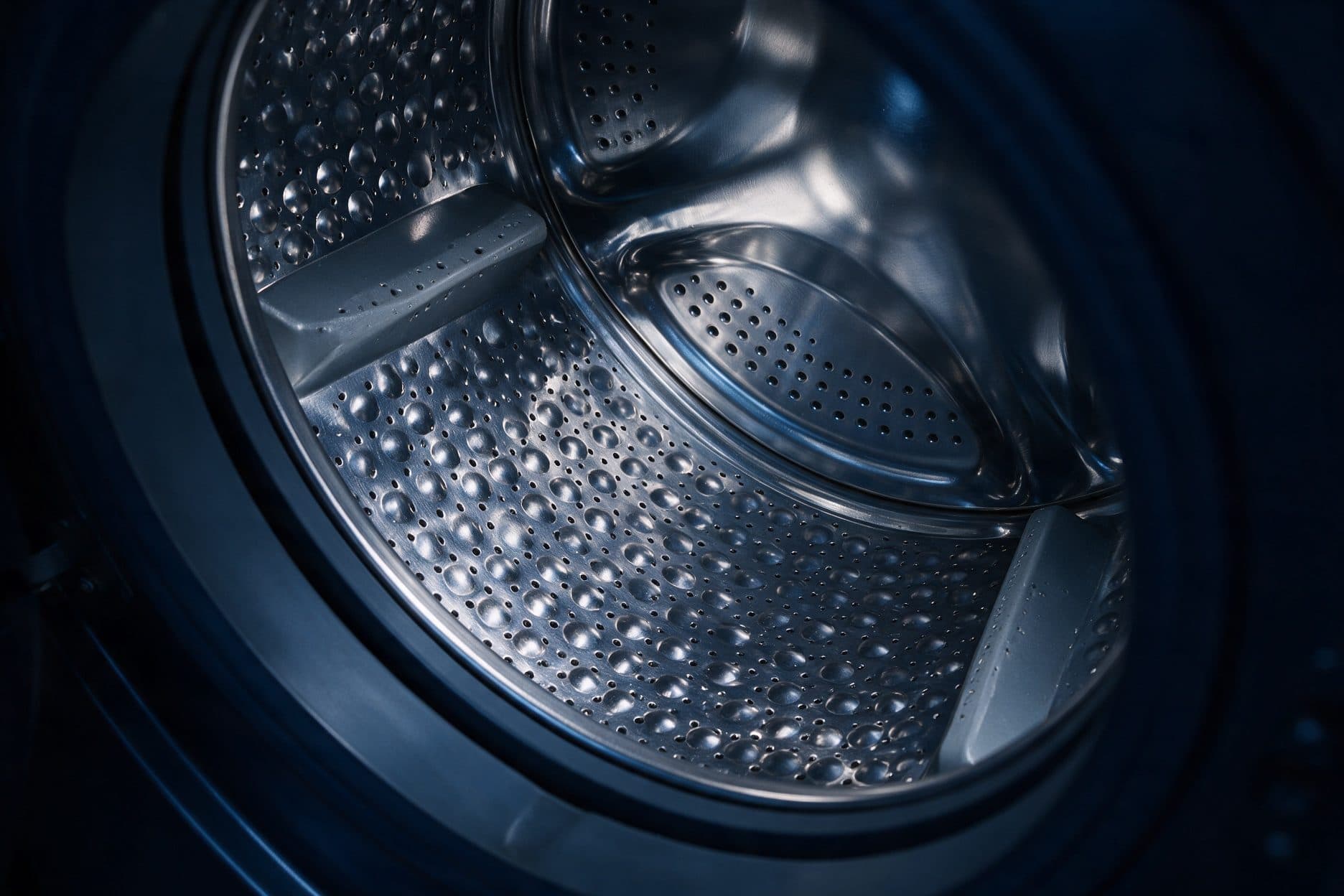 A dark navy macro of a stainless front-loader drum interior showing a raised dimpled, perforated wall raked by cool light, two wedge lifters running toward the dished perforated back wall, and a dark gasket ring at the drum mouth.
