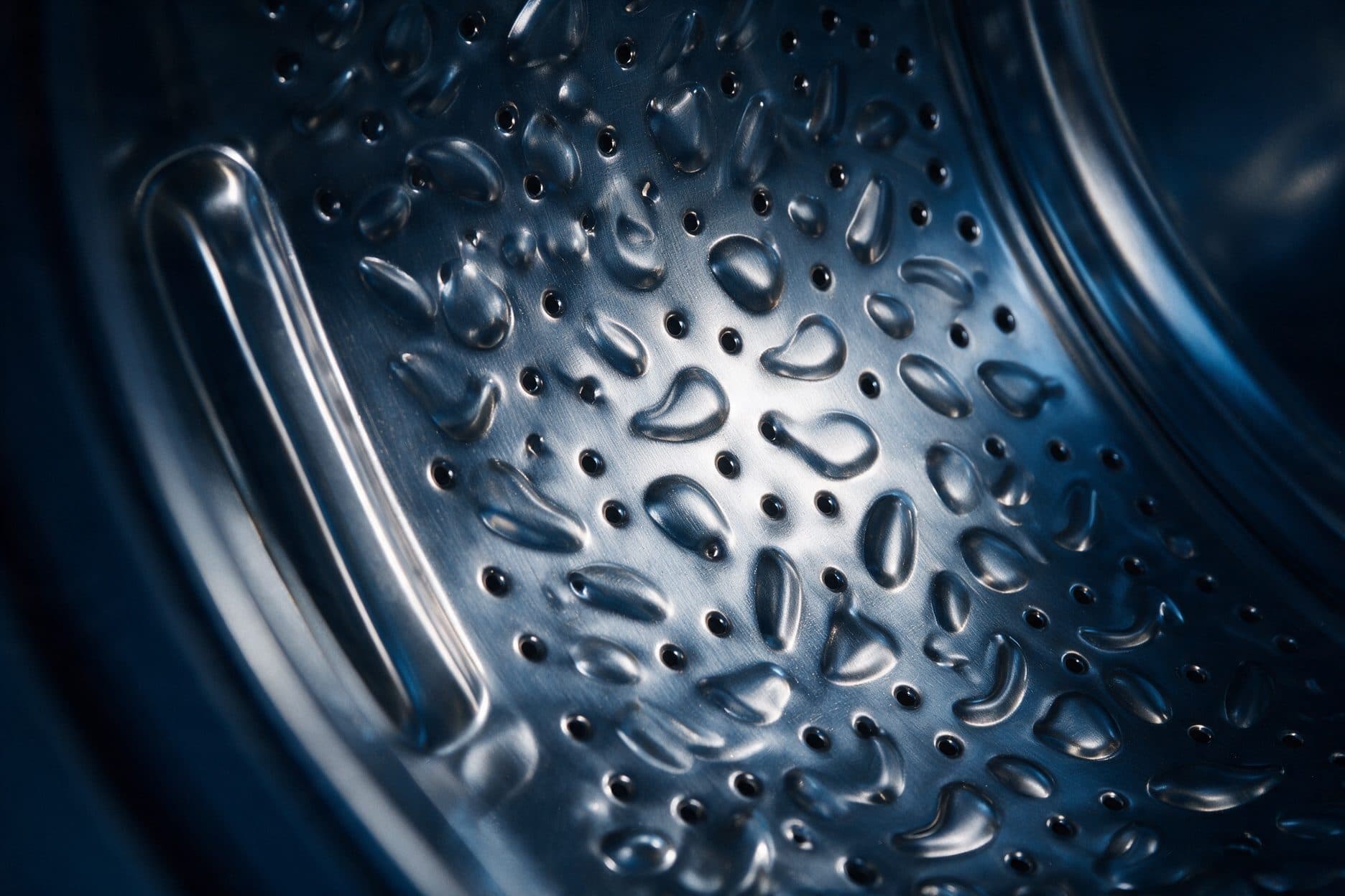 Close-up of a stainless washing-machine drum wall in blue-toned dark light with raised teardrop dimples between small drain holes and a polished lifter on the left.