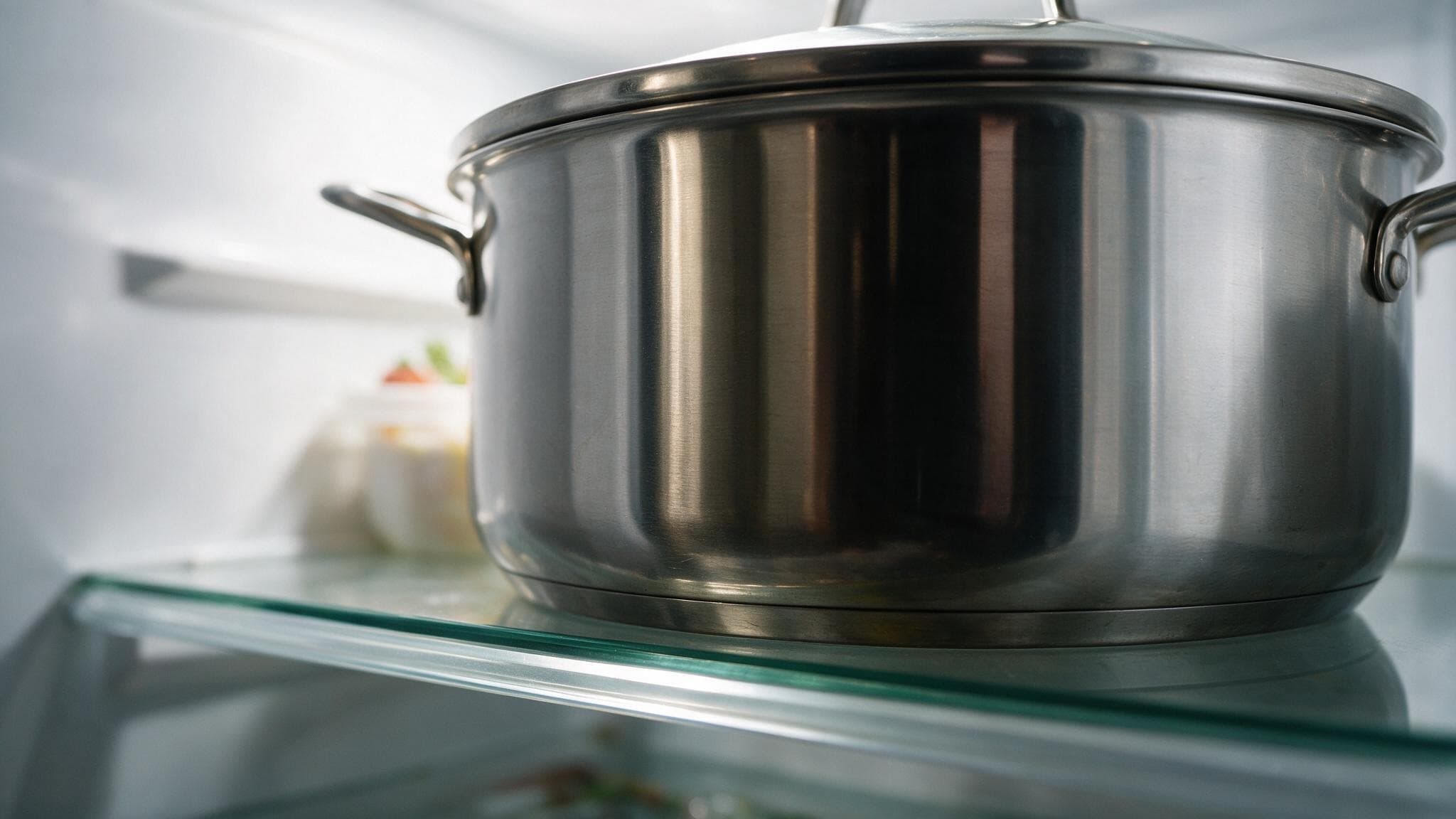 A large lidded stainless steel pot standing on a clear glass refrigerator shelf, the polished edge of the glass lit along its length.